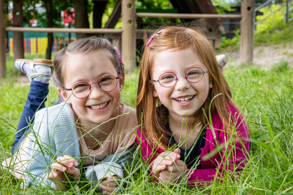 Zwei Geschwister lachen gemeinsam im Kindergarten-Garten in die Kamera.“